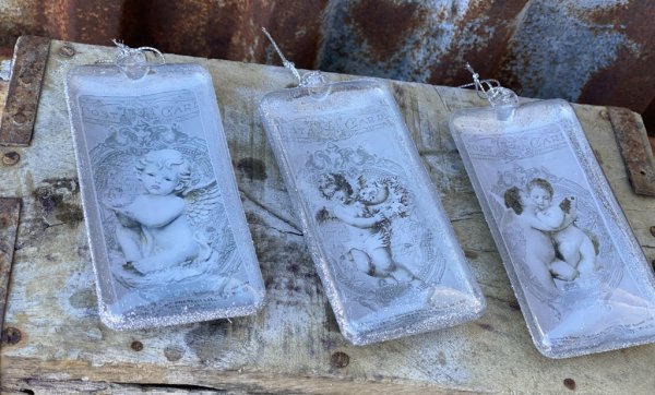 Julgranskula / dekorationskula i avlång modell med ängelmotiv. Finns i tre olika motiv -Pussande änglar -Ängel med blomsterkrans