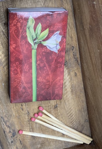 Tändstickor i vacker förpackning med motiv av en vit utslagen amaryllis på en sida och en fågel med text Happy Holiday på andra