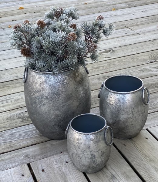 Kruka/urna/tunna med ledade handtag på sidorna. I rundad modell tillverkad i metall och går i en grå silver nyans mot antik sil