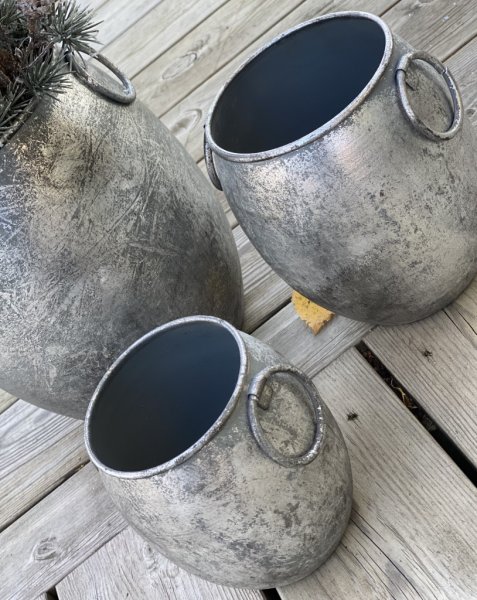 Kruka/urna/tunna med ledade handtag på sidorna. I rundad modell tillverkad i metall och går i en grå silver nyans mot antik sil